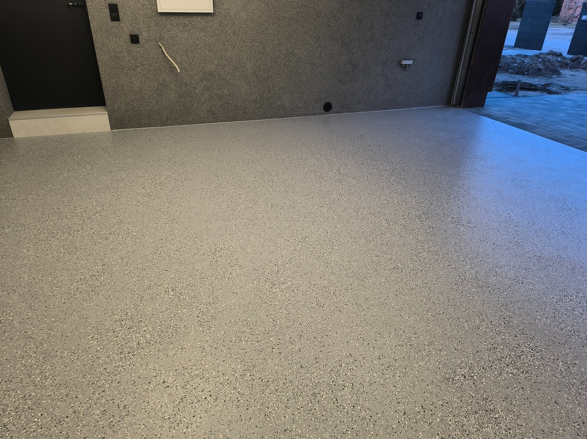 Jasnoszara posadzka żywiczna z drobnymi, ciemnymi inkluzjami, świeżo wylana w garażu. Widoczny fragment ściany z szarą strukturą i czarne drzwi po lewej.