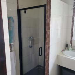 Dawid Fornalik - Wykończona łazienka z kabiną prysznicową w czarnej ramie i beżowymi płytkami z dekoracyjnym pasem. Widoczna armatura i akcesoria łazienkowe.