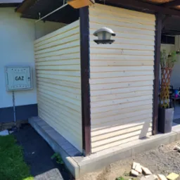 Zabudowa tarasu z desek boazeryjnych, widok z zewnątrz. Na ścianie budynku skrzynka gazowa. Elementy drewniane w kolorze naturalnym i ciemnobrązowym.