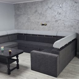 Dawid Fornalik - Szara, narożna sofa z jasnymi zagłówkami na tle ściany z efektem dekoracyjnym. Na stole filiżanka i szklanka. Widoczna podłoga z paneli.