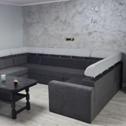 Szara, narożna sofa z jasnymi zagłówkami na tle ściany z efektem dekoracyjnym. Na stole filiżanka i szklanka. Widoczna podłoga z paneli.