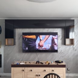 Dawid Fornalik - Szara ściana z efektem dekoracyjnym, czarne szafki wiszące i telewizor. Na komodzie dekoracje, drabina oparta o ścianę. Widok na salon po malowaniu.