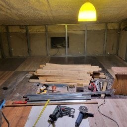 Dawid Fornalik - Poddasze w trakcie adaptacji: metalowa konstrukcja, izolacja, narzędzia, deski, lampa. Widoczne etapy prac przygotowawczych do wykończenia wnętrza.