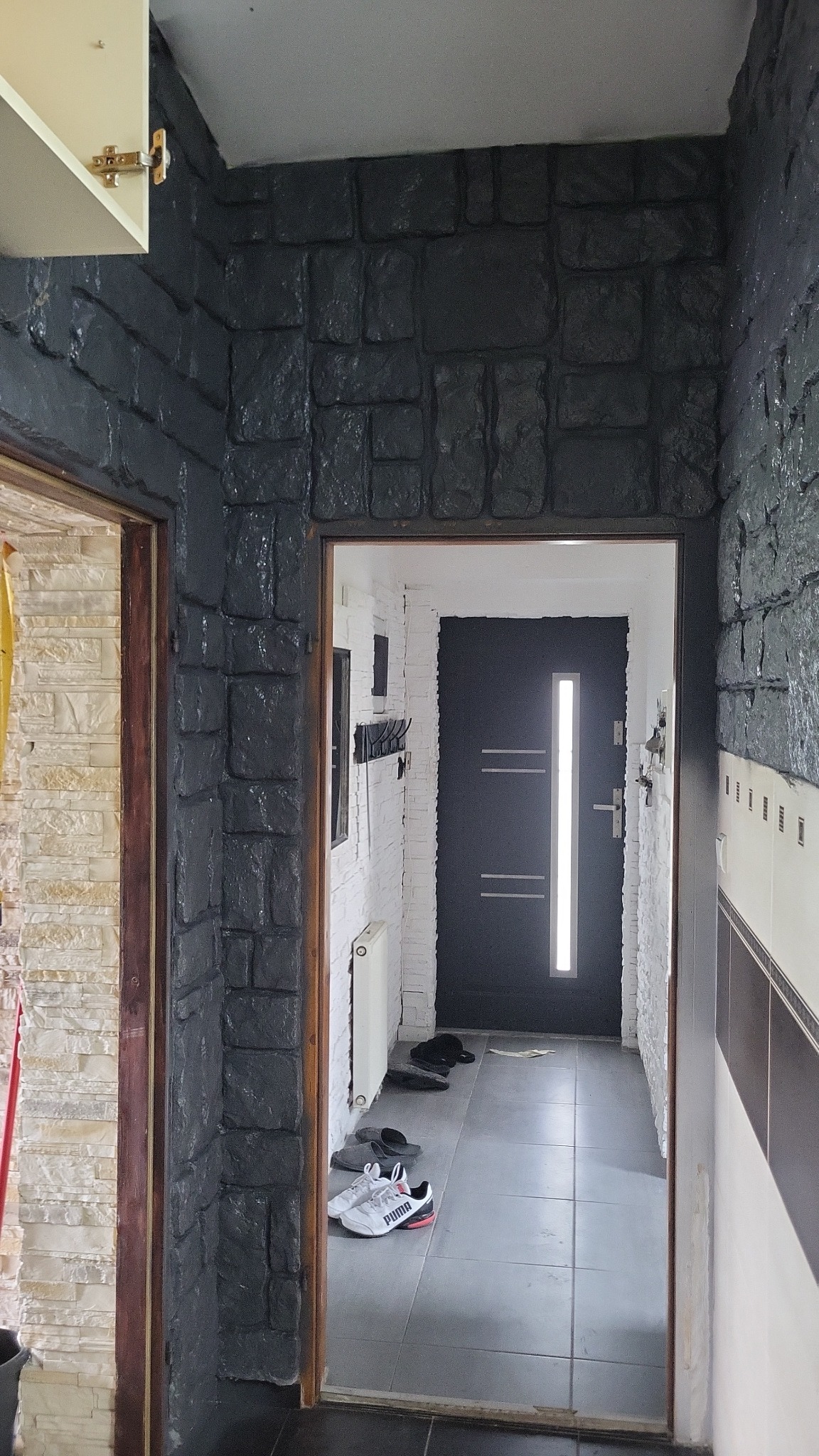 Korytarz z czarną, kamienną okładziną ścian i widokiem na białe wnętrze z drzwiami wejściowymi. Buty leżą na podłodze. Kontrast faktur i kolorów.