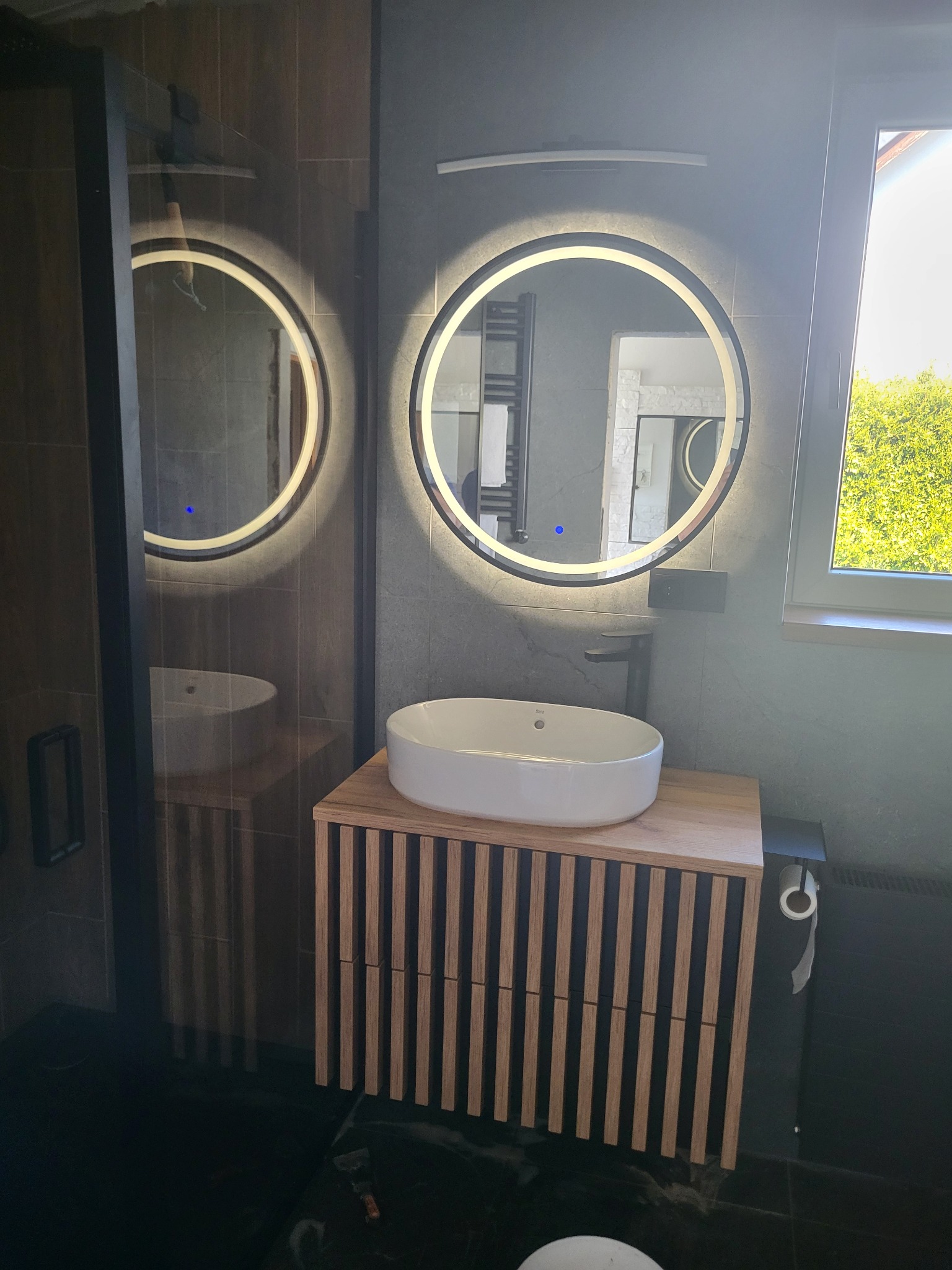 Nowoczesna łazienka po renowacji w Zebrzydowicach. Okrągłe lustra z podświetleniem LED, drewniana szafka pod umywalką, ciemne płytki. Minimalistyczny design.