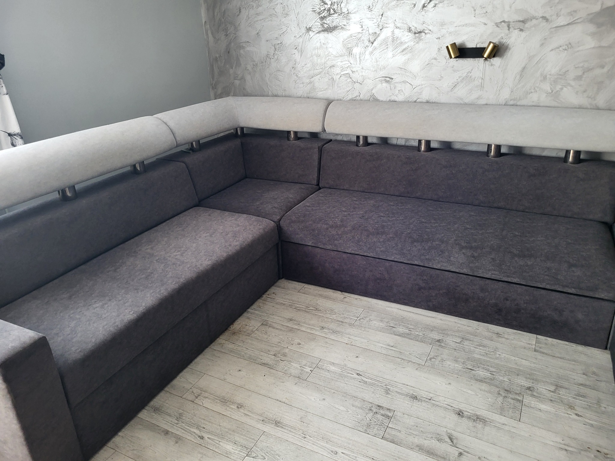 Szary narożnik z jasnymi zagłówkami i metalowymi nóżkami na tle ściany z efektem betonu i złotym oświetleniem. Widok na drewnopodobną podłogę.