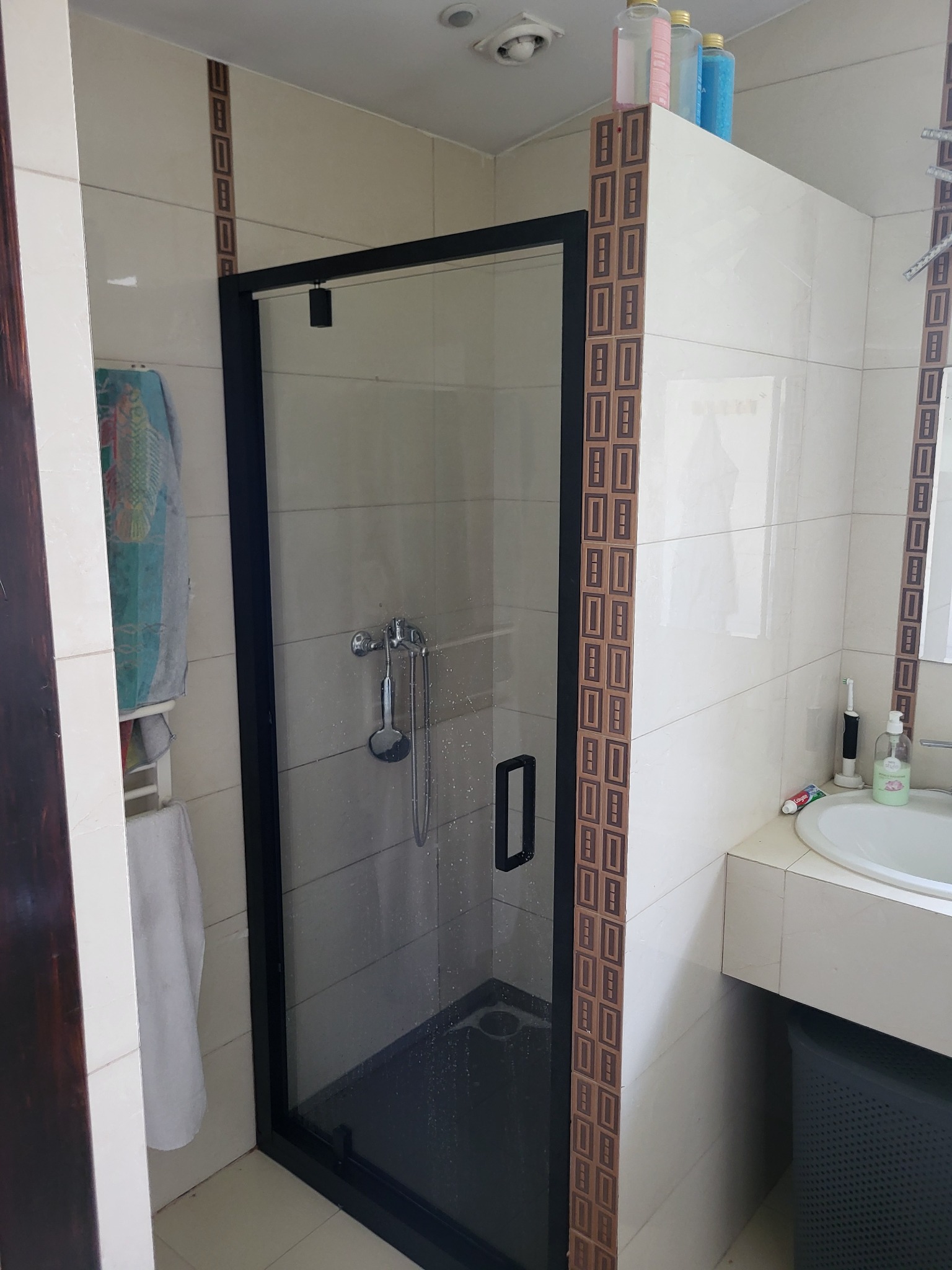 Wykończona łazienka z kabiną prysznicową w czarnej ramie i beżowymi płytkami z dekoracyjnym pasem. Widoczna armatura i akcesoria łazienkowe.