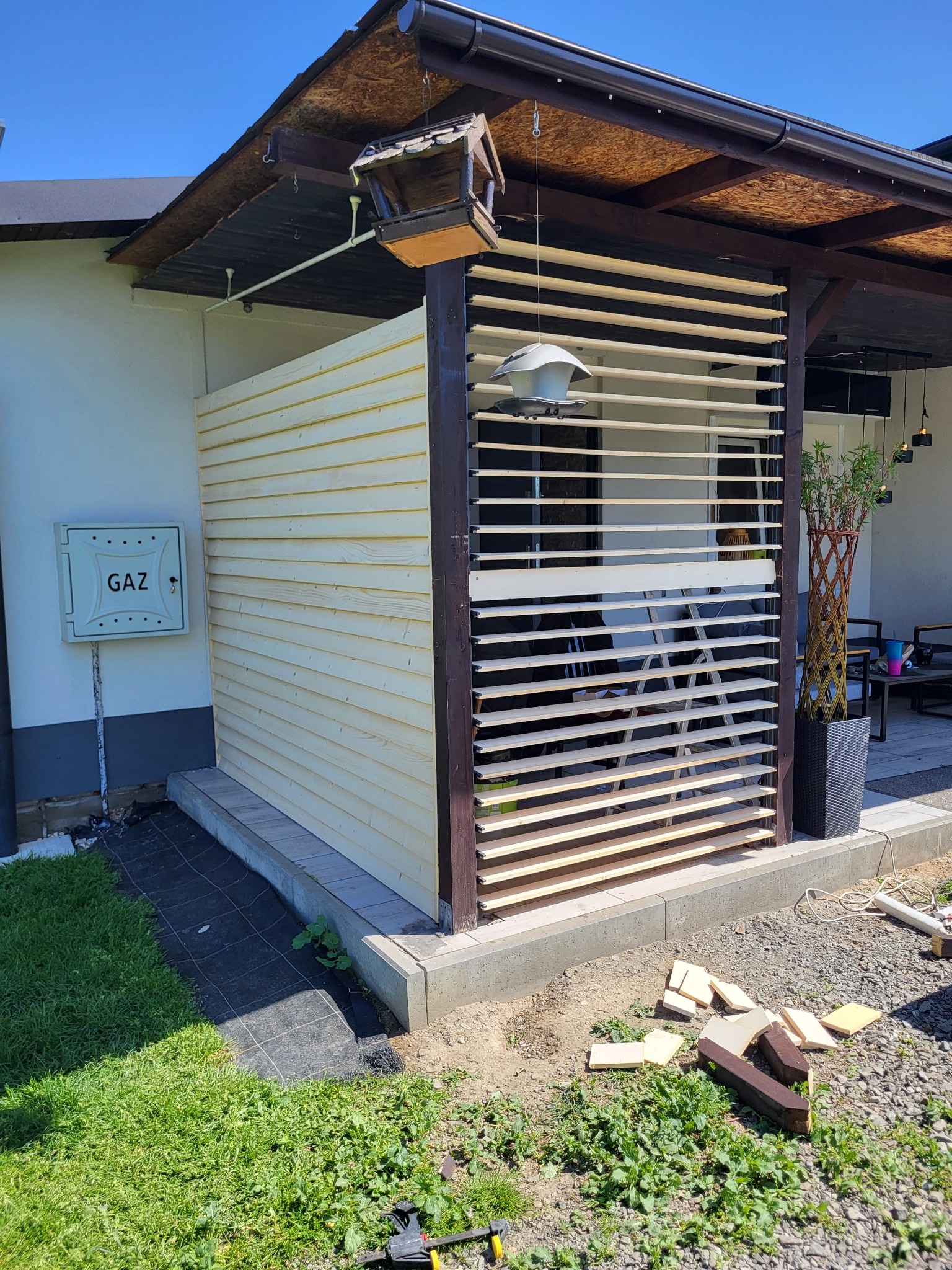 Drewniana konstrukcja tarasu w trakcie budowy, z widocznymi elementami szkieletu i listwami, narzędzia na ziemi, skrzynka gazowa na ścianie budynku.