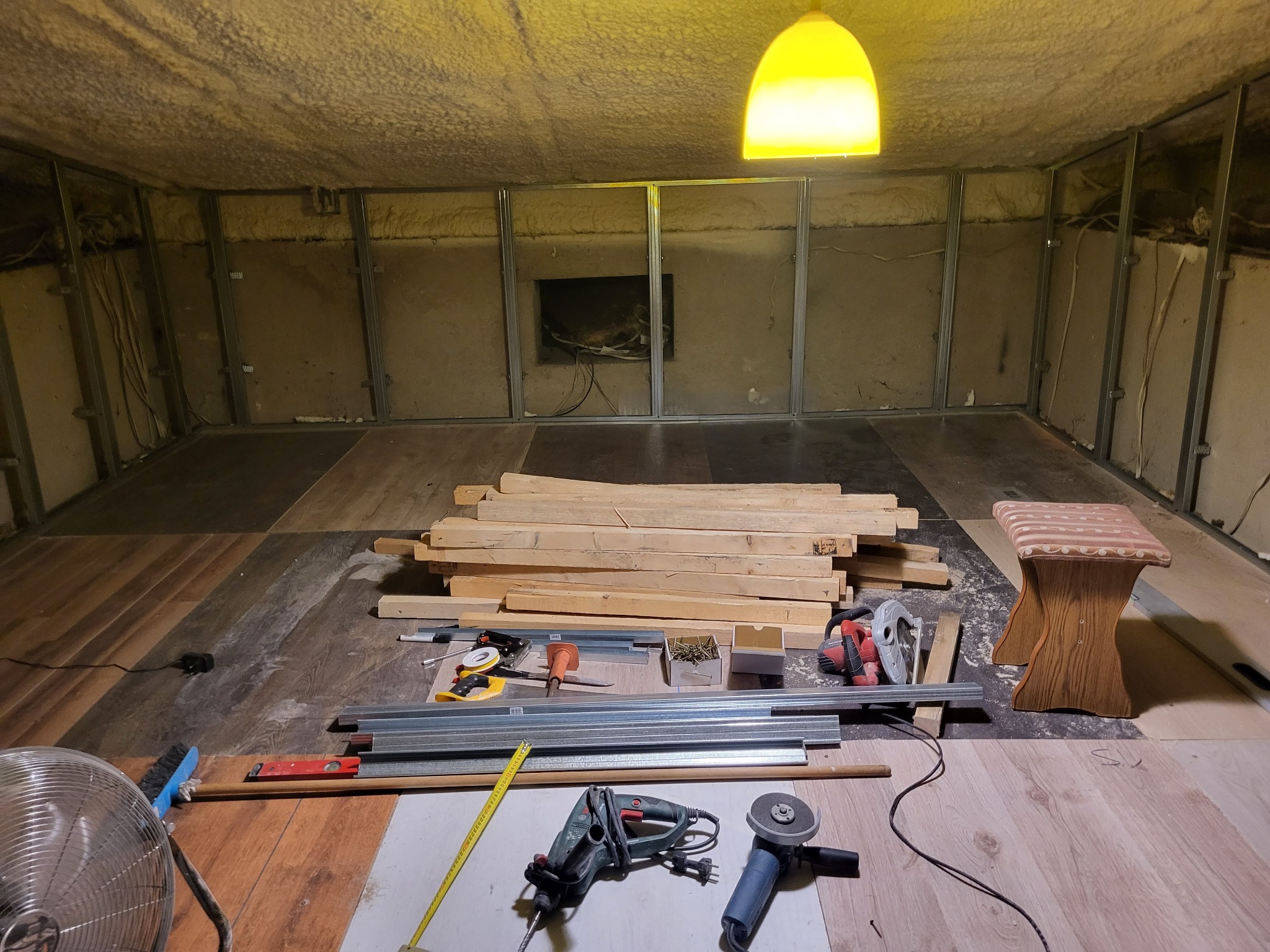 Poddasze w trakcie adaptacji: metalowa konstrukcja, izolacja, narzędzia, deski, lampa. Widoczne etapy prac przygotowawczych do wykończenia wnętrza.