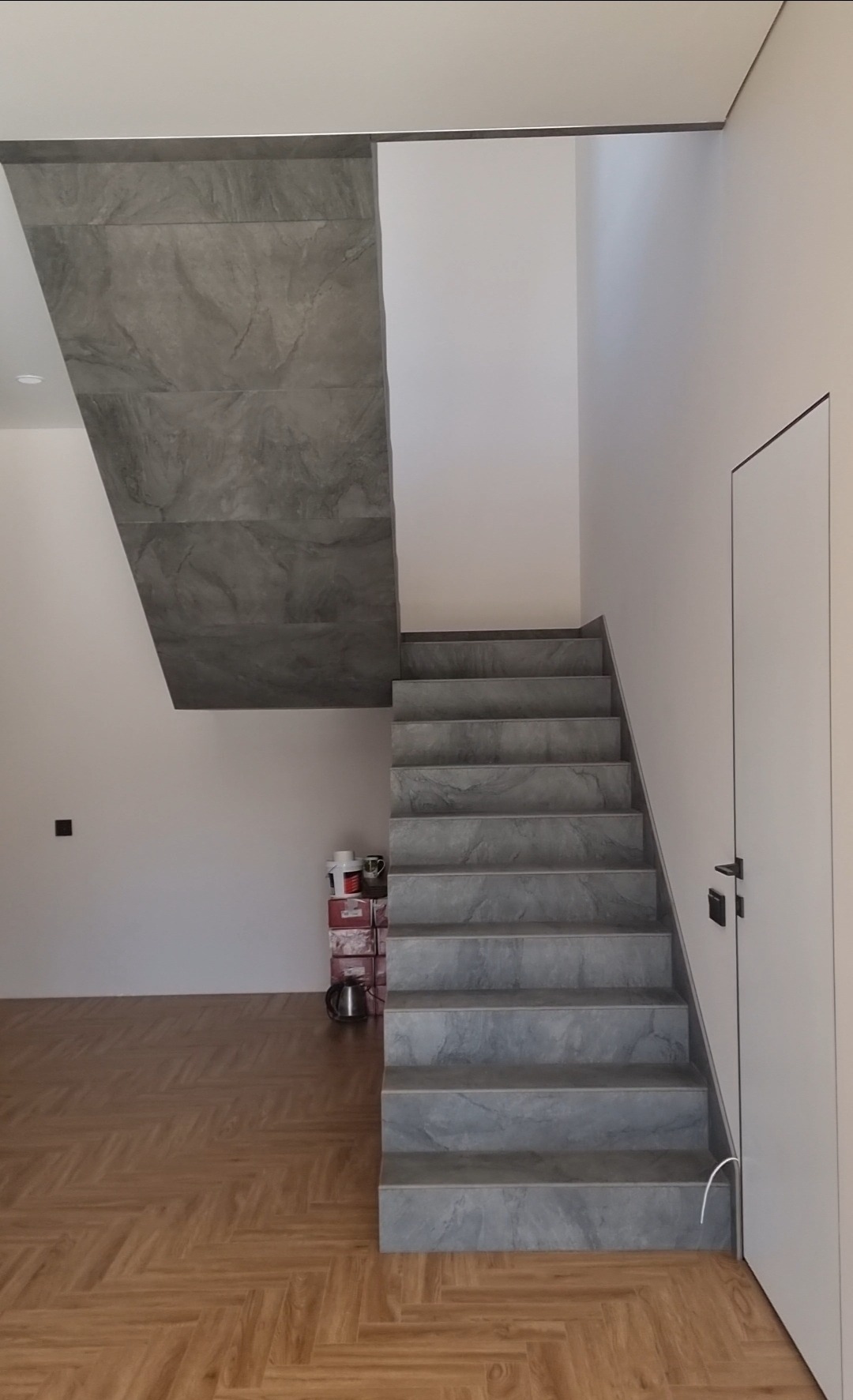 Wykończone schody z szarych płytek imitujących kamień, prowadzące na piętro. Na podłodze parkiet ułożony we wzór jodełki. Minimalistyczny design.