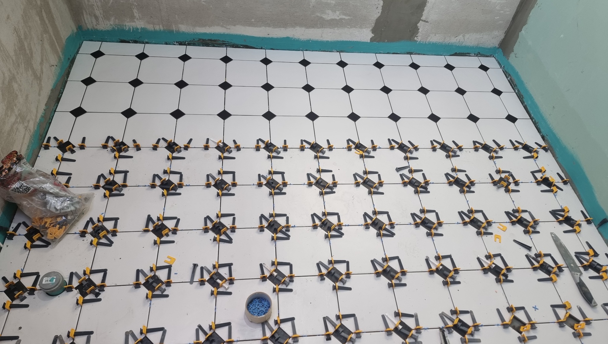 Białe płytki podłogowe z czarnymi akcentami, częściowo ułożone, z systemem poziomowania i narzędziami w tle. Widok z góry na wnętrze w trakcie wykańczania.