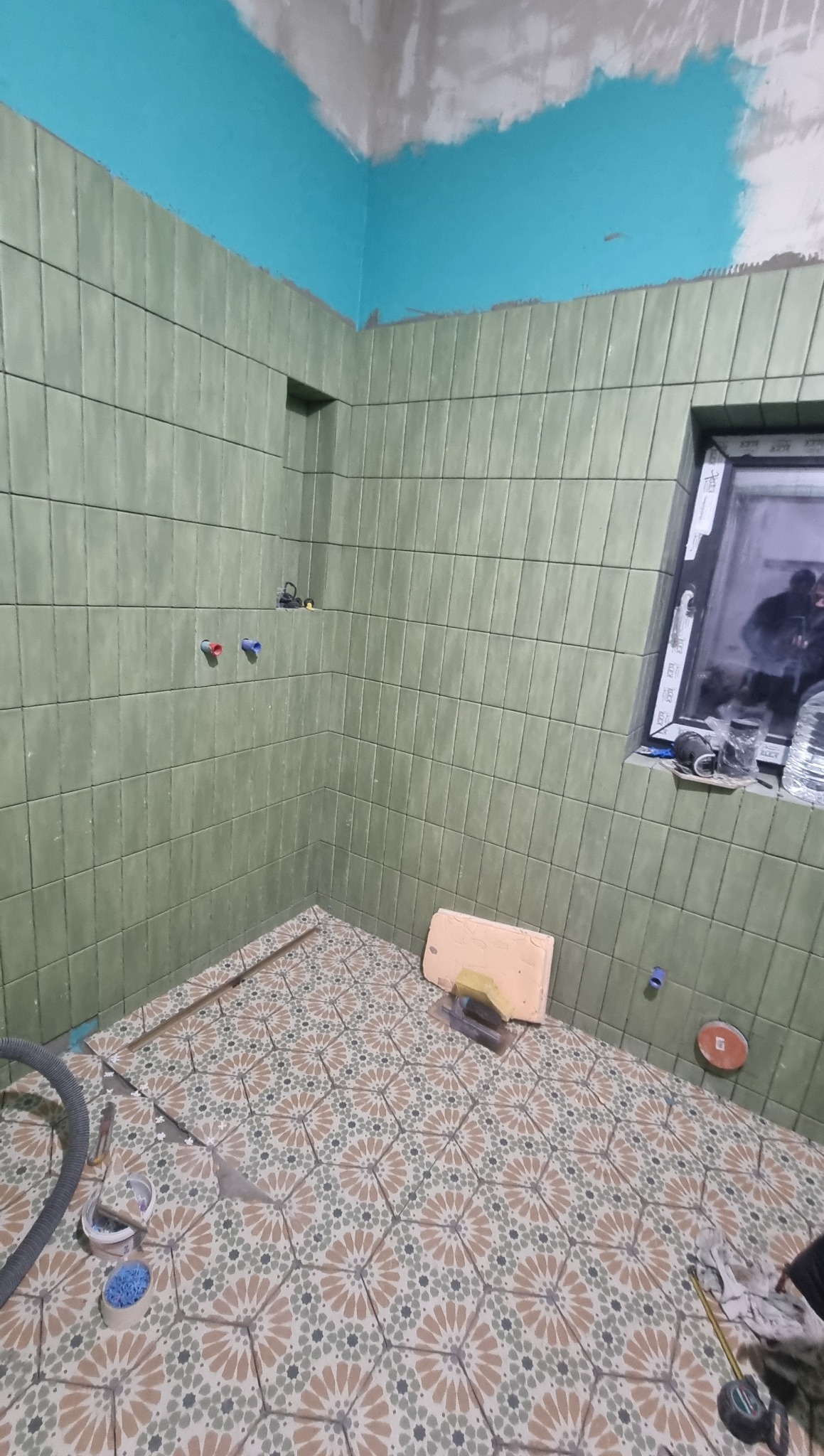 Wnętrze łazienki w trakcie prac. Ściany wyłożone zielonymi płytkami, podłoga w kwiatowy wzór. Widoczne narzędzia i akcesoria glazurnicze. Częściowo pomalowany sufit na niebiesko.