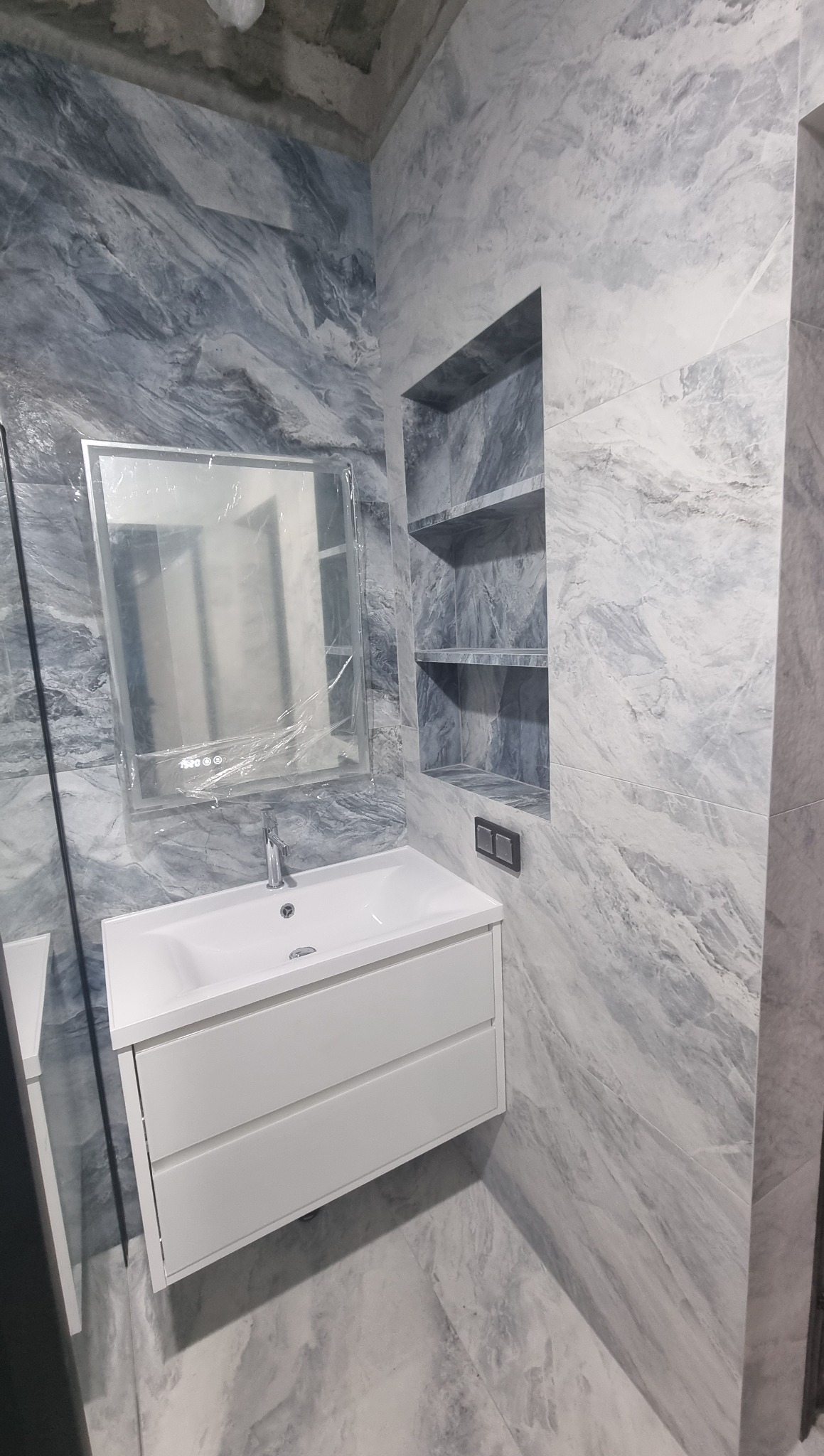 Wykończona łazienka z szarymi płytkami imitującymi marmur. Umywalka z szafką w kolorze białym i wnęka z półkami w ścianie. Lustro jeszcze w folii ochronnej.