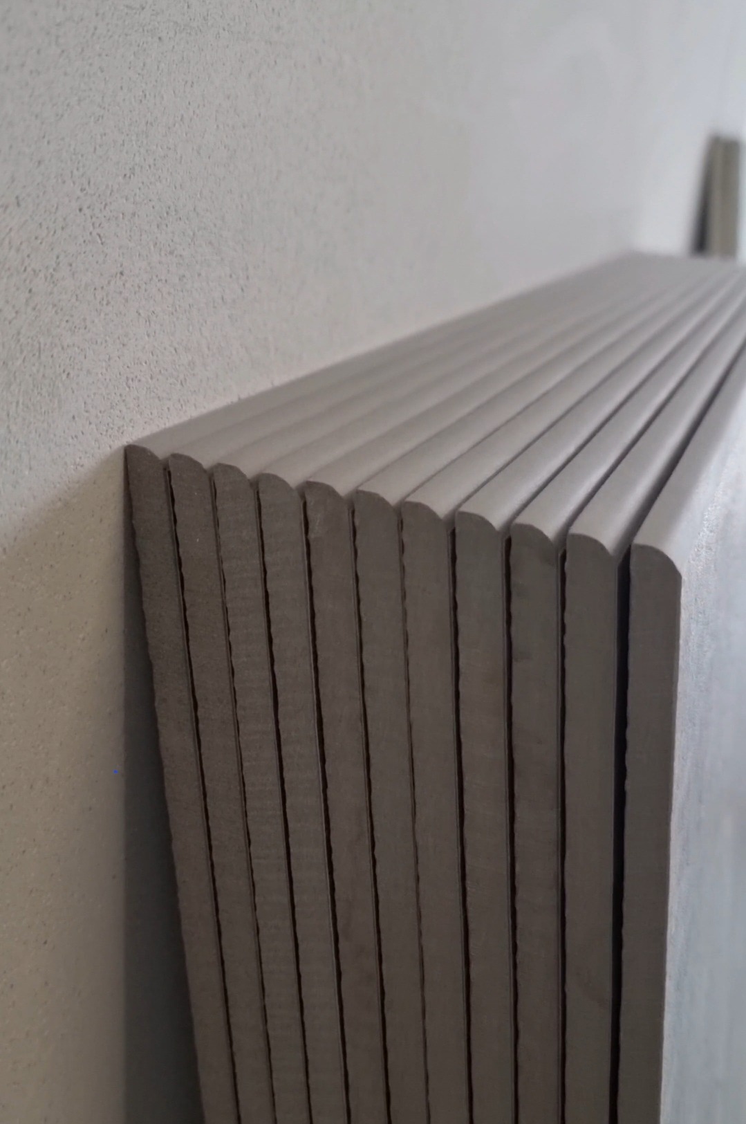 Ułożone pionowo, szare płyty gipsowe z zaokrąglonymi krawędziami, opierają się o szorstką, jasną ścianę. Widok z boku, w tle fragment kolejnej płyty.