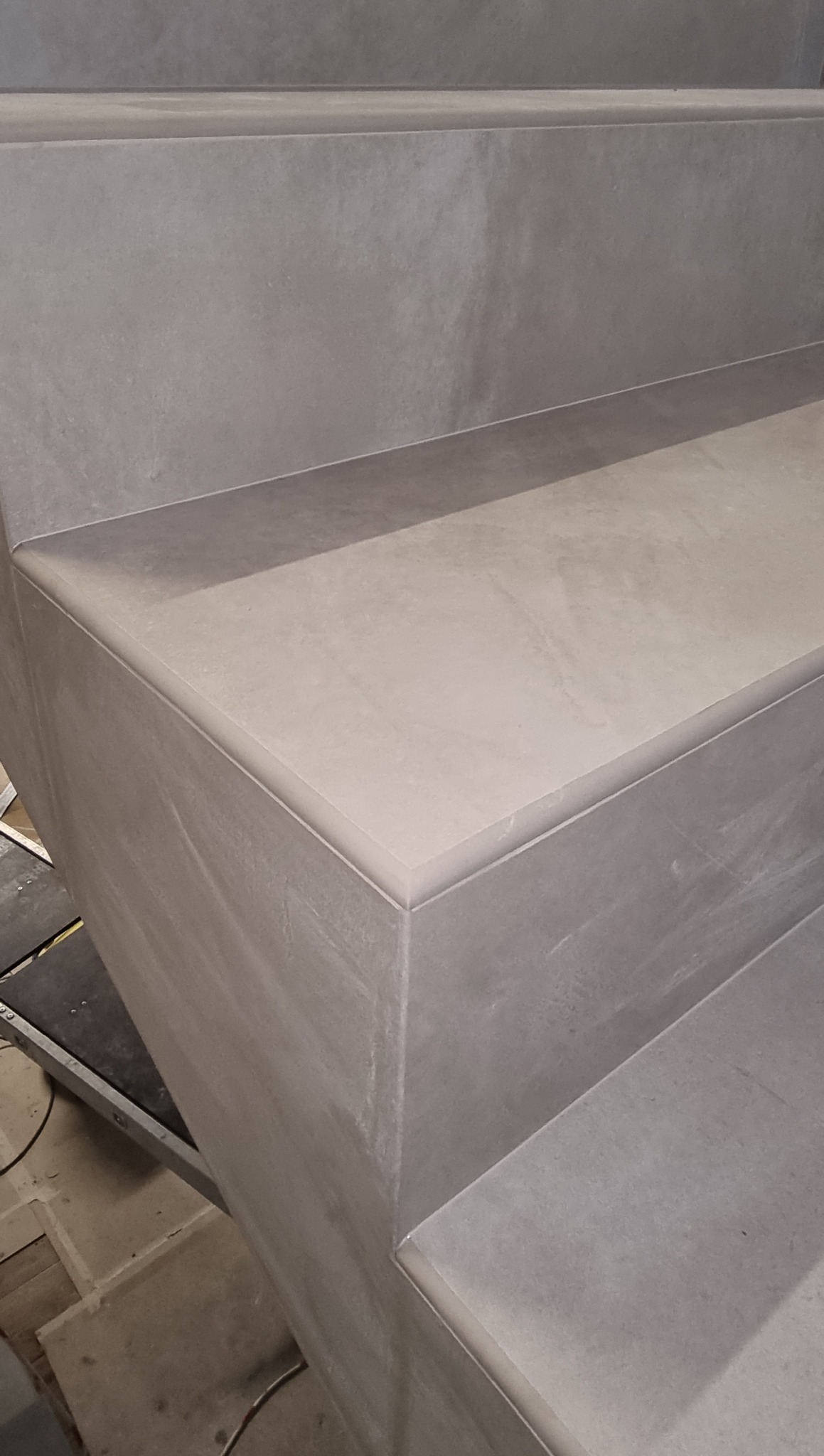 Wykończone schody z szarych, matowych płytek ceramicznych z zaokrąglonymi krawędziami. Detal narożnika stopnia, widoczne fugi. Estetyczne i staranne wykonanie.