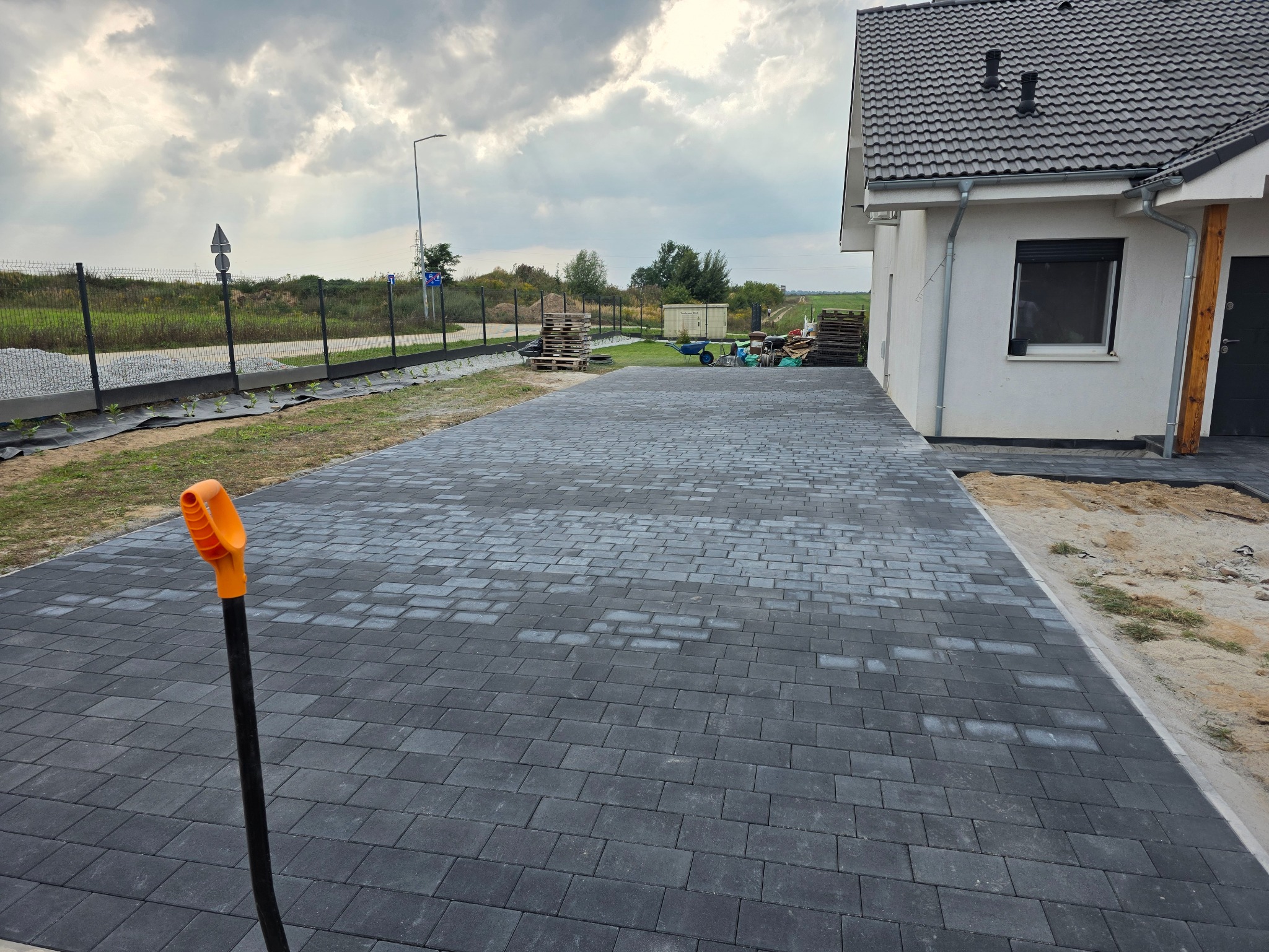 Ułożona, szara kostka brukowa prowadząca do domu jednorodzinnego. W tle widoczny teren działki i ogrodzenie. Na pierwszym planie wbita łopata.