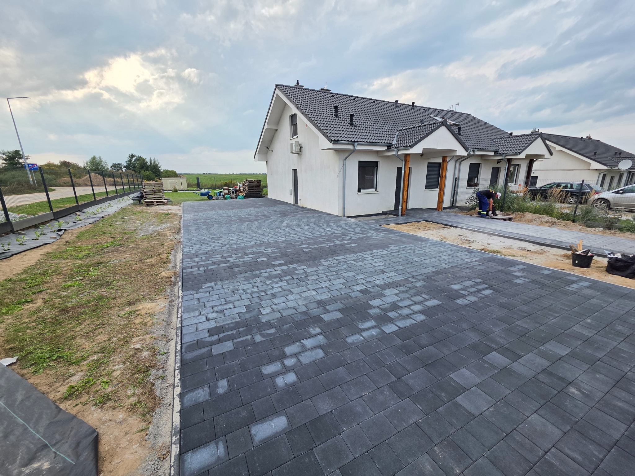 Ułożona, grafitowa kostka brukowa prowadząca do białego domu z ciemną dachówką. W tle widoczne prace wykończeniowe i ogrodzenie.