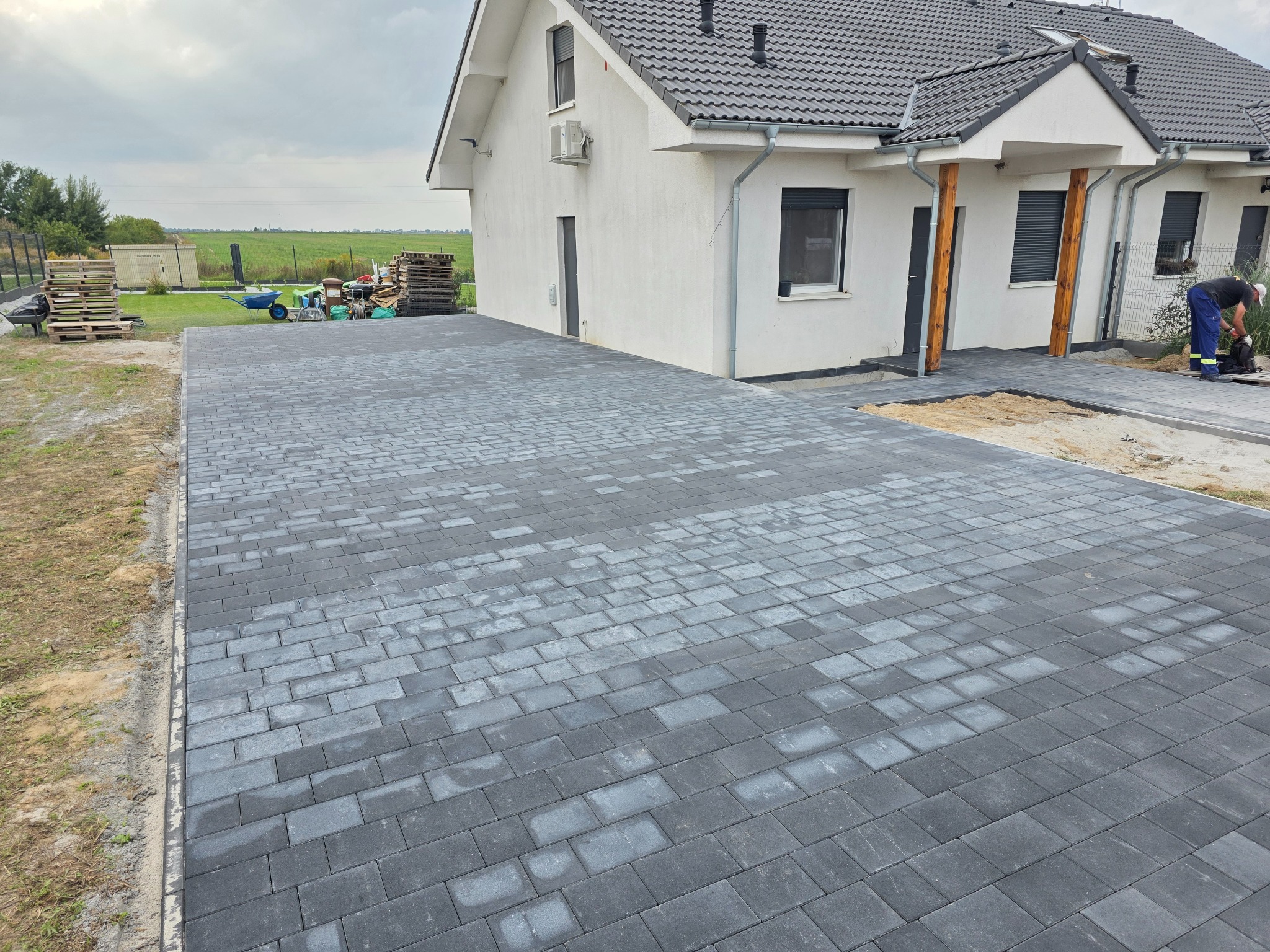Ułożona, szara kostka brukowa na podjeździe przed domem jednorodzinnym. W tle widoczne palety i narzędzia, a także pracownik podczas wykonywania prac wykończeniowych.