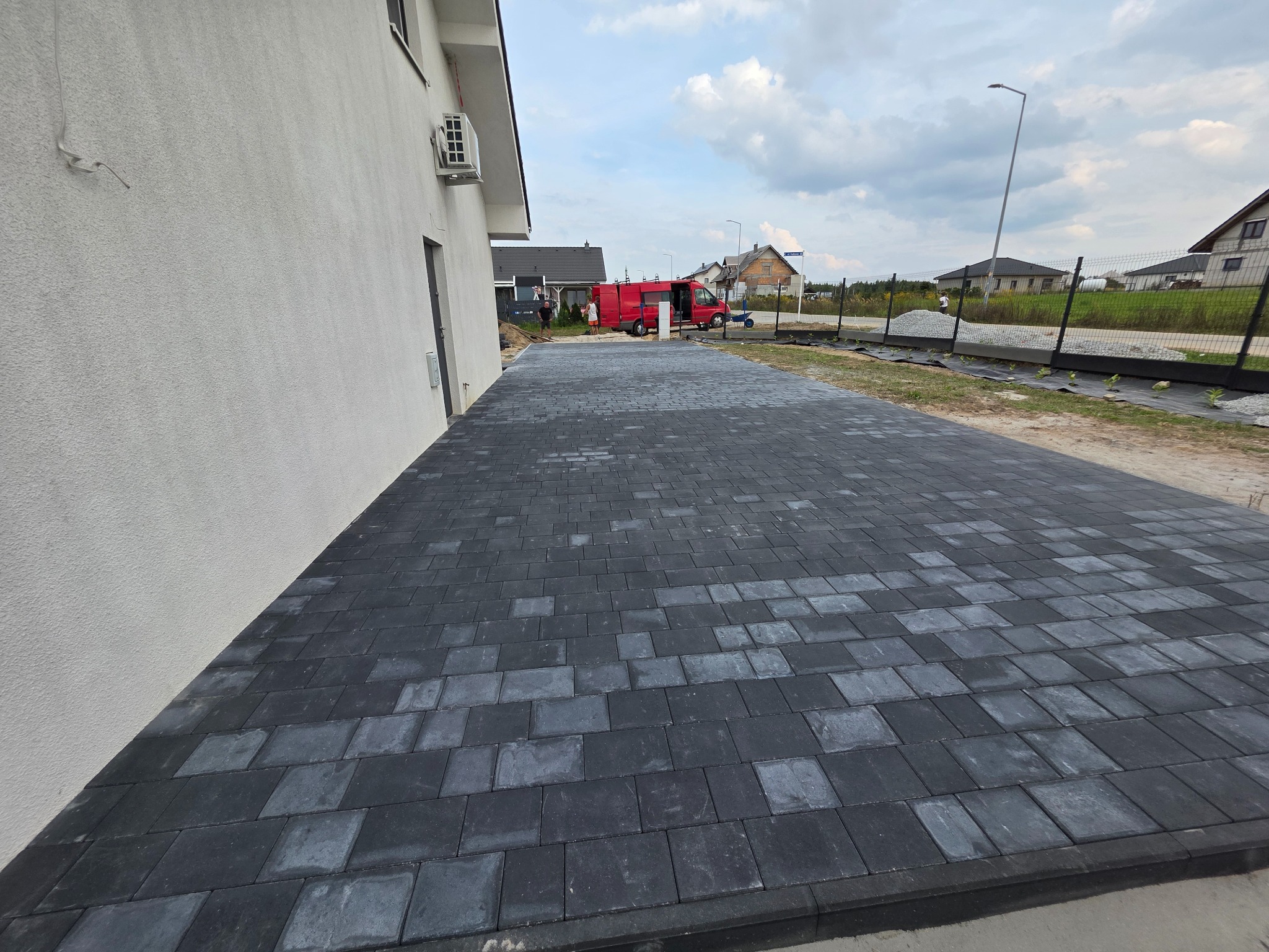 Ułożona, czarna kostka brukowa przy ścianie domu. Widoczny fragment ogrodzenia i budynki w tle. Kostka ułożona w regularny wzór, tworząc estetyczny podjazd.