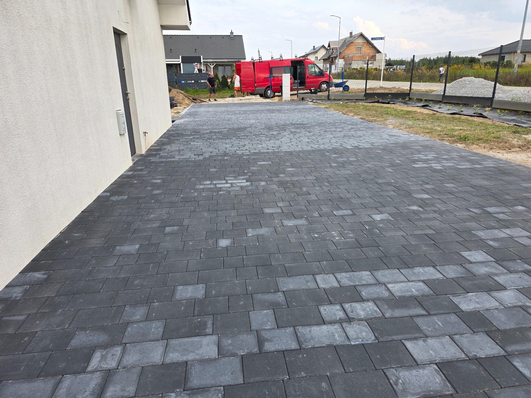 Ułożona, szara kostka brukowa na podjeździe domu jednorodzinnego. W tle czerwony bus i prace przygotowawcze do zagospodarowania terenu zielonego.