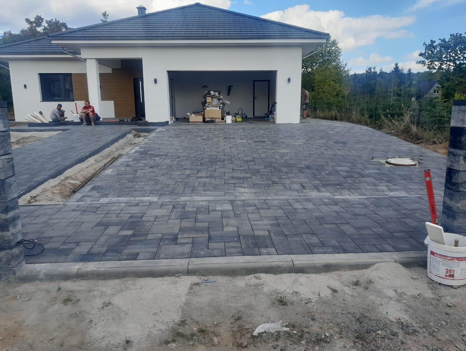 Nowoczesny podjazd z szarej kostki brukowej przed domem z garażem. Częściowo ułożona kostka, widoczne obrzeża i materiały budowlane. Prace brukarskie w toku.
