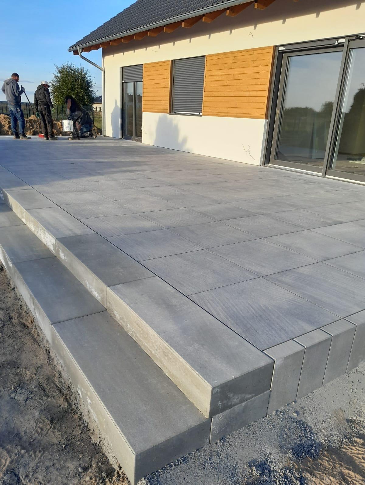 Nowoczesny taras z szarych płyt betonowych i schodami, w tle dom z drewnianą elewacją. Trzech robotników w oddali, prace wykończeniowe.