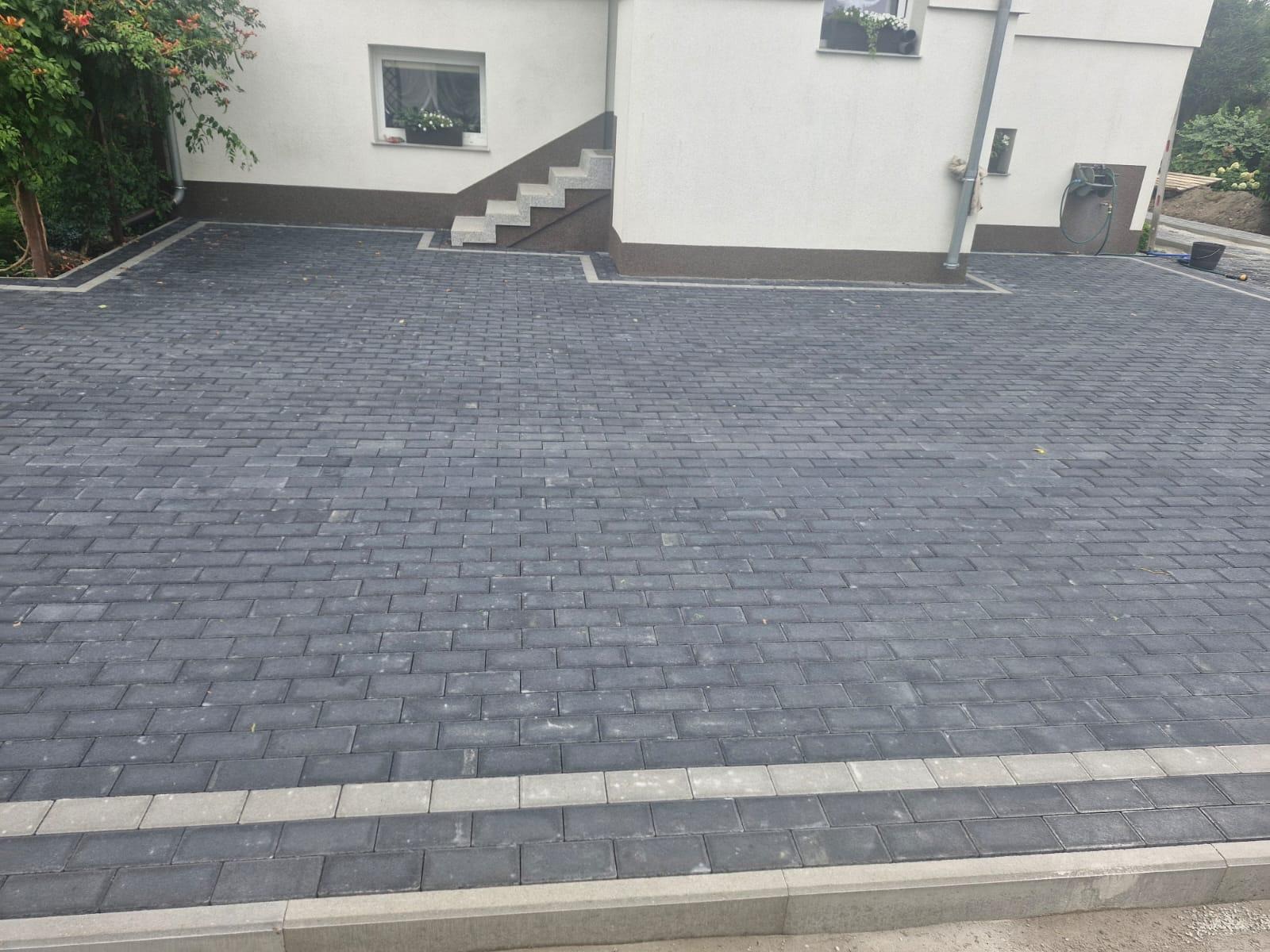 Ułożona, szara kostka brukowa na podjeździe domu z białymi ścianami i schodami wejściowymi. Obramowanie posesji z jasnych kostek. Widok z góry, bez osób.