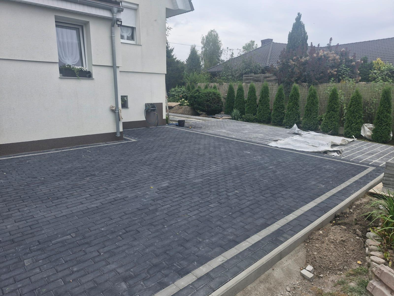 Ułożona, grafitowa kostka brukowa przed domem z jasną elewacją. Widoczne obrzeża i fragment dalszej części posesji z tujami. Częściowo przykryta kostka w tle.