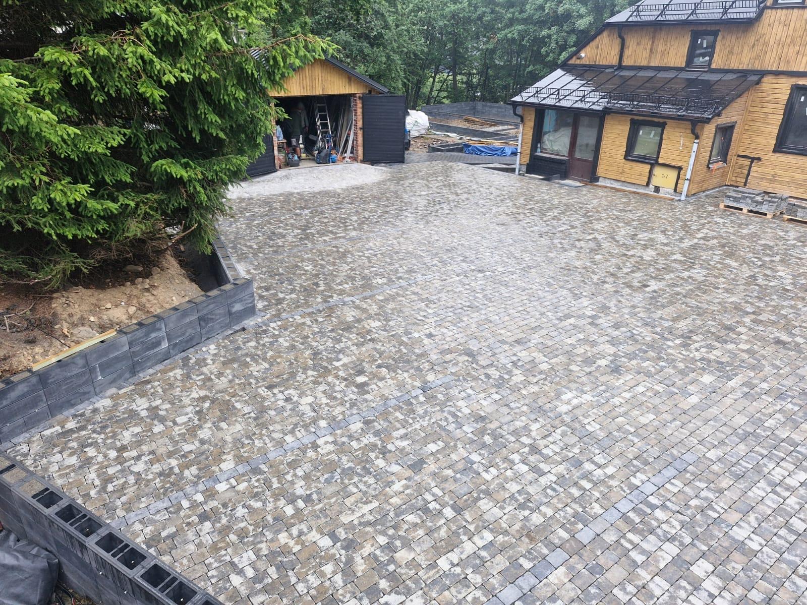 Gotowa nawierzchnia z kostki brukowej w Janczewie, widok z góry. Szara kostka ułożona na posesji z budynkami i zielenią w tle. Kostka w odcieniach szarości i beżu.