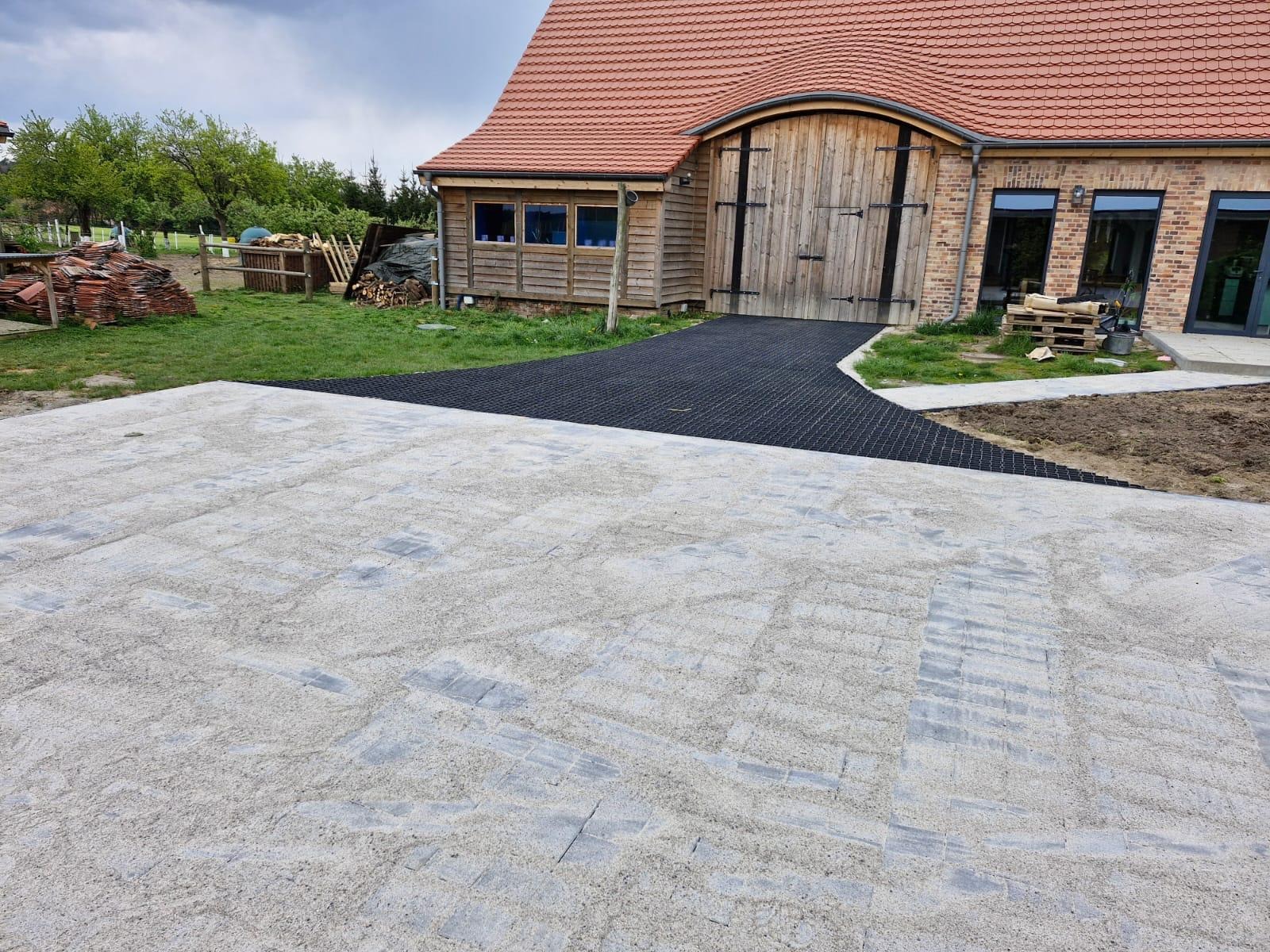 Układanie przepuszczalnej nawierzchni z kostki brukowej i kratki. Widok na dom z drewnianymi drzwiami i fragmentem ogrodu. Realizacja w toku - piasek na kostce.