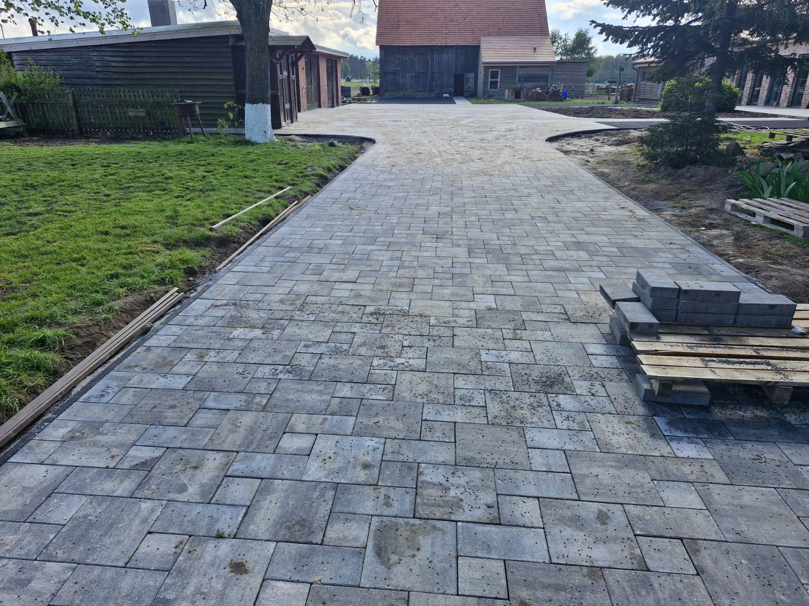 Ułożona szara kostka brukowa tworzy ścieżkę na posesji. W tle budynki gospodarcze i zieleń. Paleta z kostką obok ścieżki. Widok na ułożoną nawierzchnię.