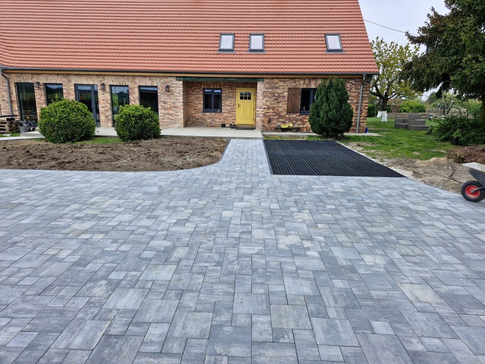 Ułożona szara kostka brukowa przed domem z żółtymi drzwiami i fragmentem trawnika z krzewami. Widoczna ażurowa nawierzchnia. Słoneczny dzień.