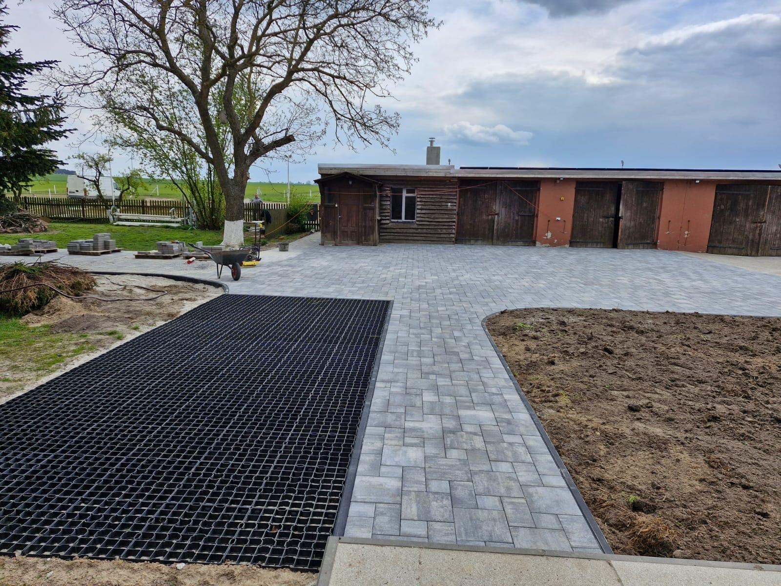 Ułożona szara kostka brukowa tworząca ścieżkę i plac. Po lewej stronie widoczna czarna kratka trawnikowa, a w tle budynek gospodarczy i drzewo.