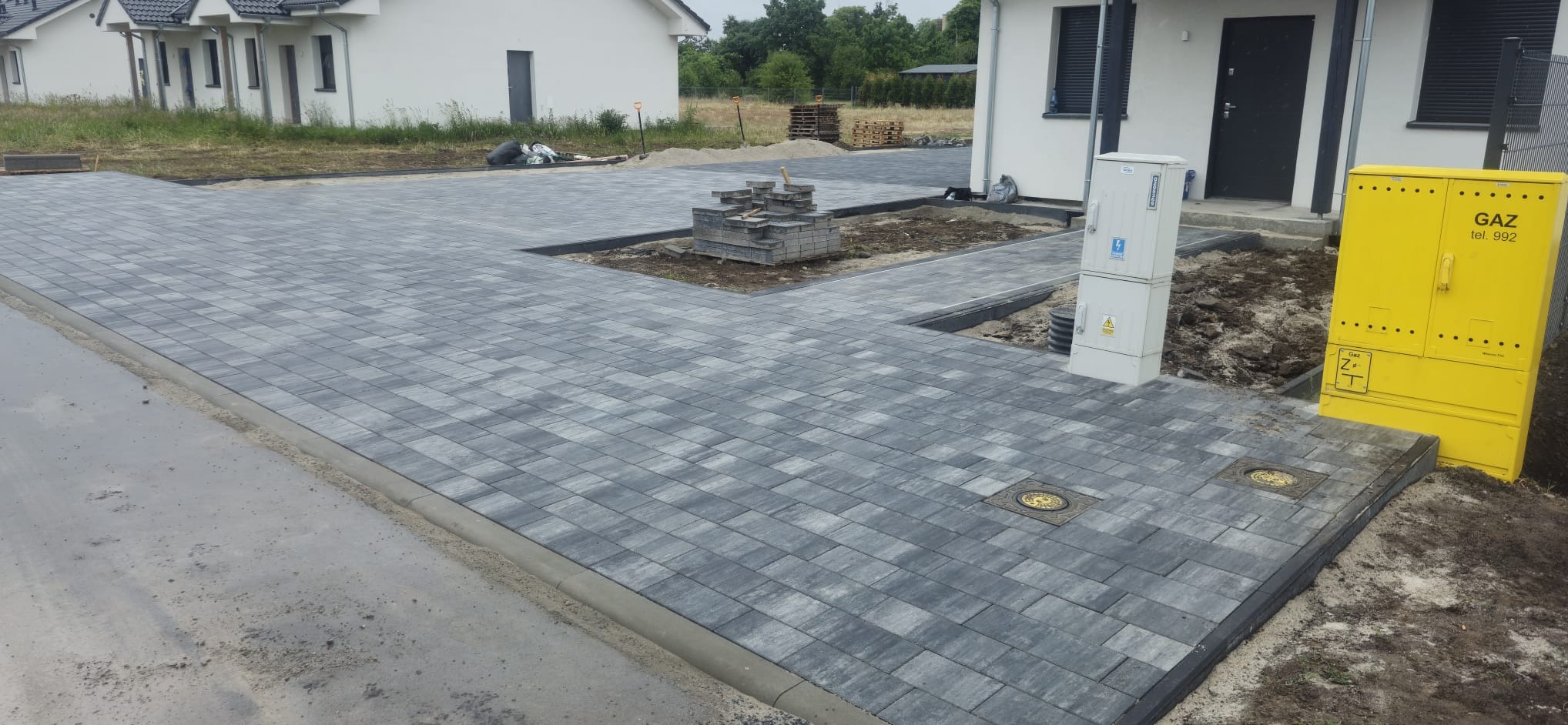 Szara kostka brukowa ułożona na podjeździe przed domem, widoczne studzienki i żółta skrzynka gazowa. Częściowo ułożona kostka, przygotowana do dalszych prac.