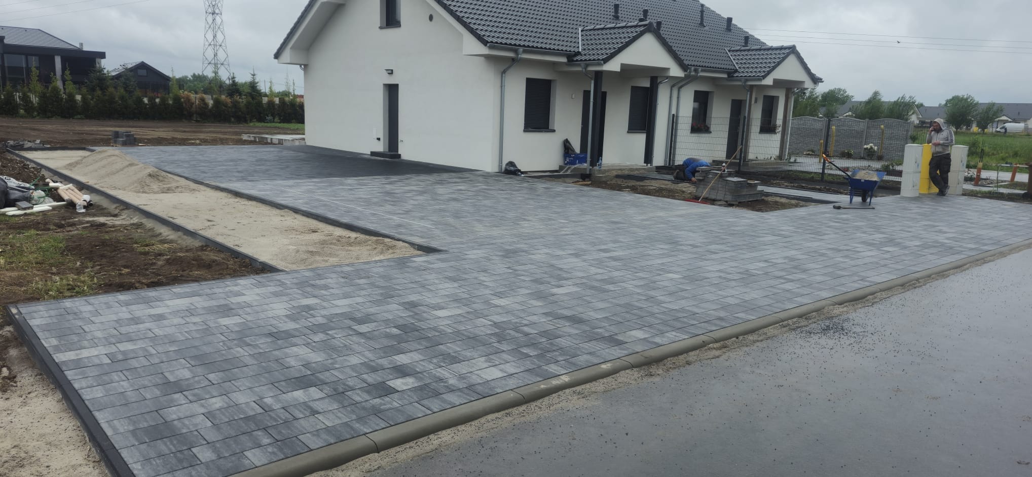 Ułożona szara kostka brukowa na podjeździe domu jednorodzinnego. Część powierzchni jeszcze nieukończona, widoczny piasek i materiały budowlane. Prace brukarskie w toku.