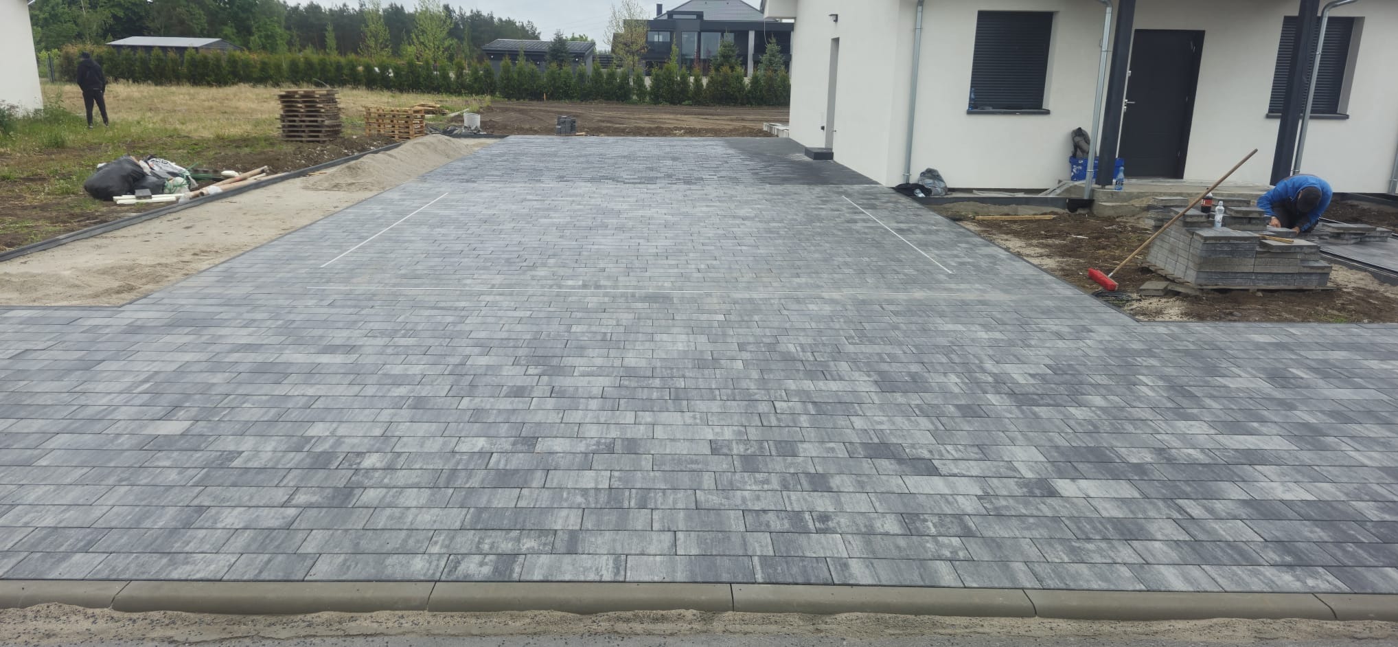 Częściowo ułożona, szara kostka brukowa na podjeździe domu jednorodzinnego. W tle osoba i palety. Na pierwszym planie obrzeże. Prace brukarskie w toku.