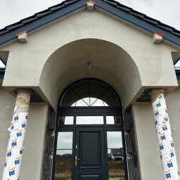 VisionTherm - Wejście do domu z czarnymi drzwiami i łukowym portalem w trakcie wykańczania elewacji. Kolumny zabezpieczone folią. Widoczne detale architektoniczne i surowy stan prac.
