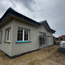 VisionTherm - Dom jednorodzinny w trakcie ocieplania elewacji, widoczne rusztowania i materiały budowlane. Szara elewacja, białe okna, pochmurne niebo w tle.