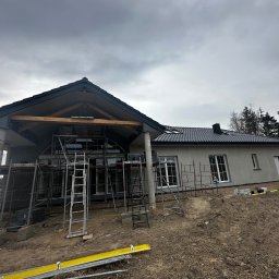 VisionTherm - Dom w trakcie budowy z szarym tynkiem, widoczne rusztowania i elementy konstrukcyjne dachu. Pochmurne niebo w tle. Ujęcie z dołu.