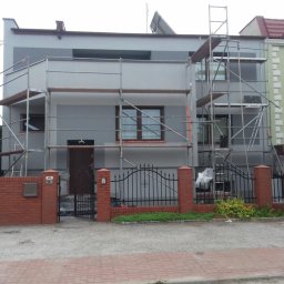 VisionTherm - Dom jednorodzinny w trakcie malowania elewacji, otoczony rusztowaniami. Widoczny fragment ogrodzenia z cegły i metalu. Prace w toku, szara kolorystyka budynku.