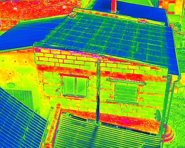 Termowizyjne zdjęcie budynku z bliska, ukazujące rozkład temperatur na ścianach i dachu. Widoczne okna i fragmenty konstrukcji. Nietypowa kolorystyka.