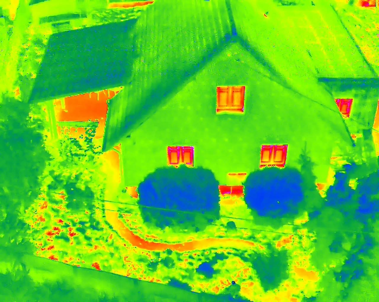Termowizja domu jednorodzinnego z ogrodem, ukazująca straty ciepła przez okna i dach. Zielono-pomarańczowa kolorystyka termogramu.