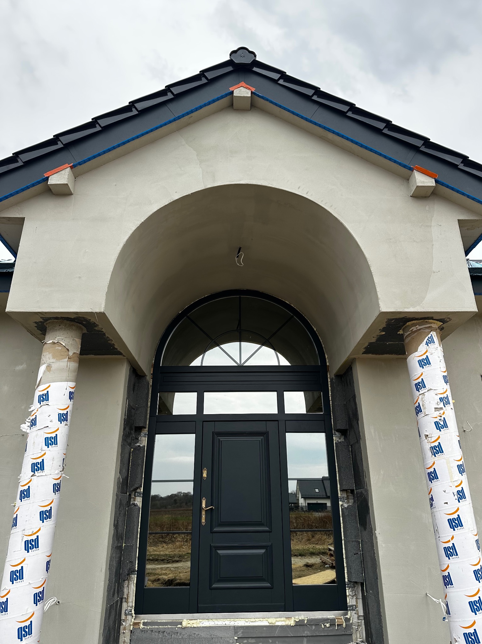 Wejście do domu z czarnymi drzwiami i łukowym portalem w trakcie wykańczania elewacji. Kolumny zabezpieczone folią. Widoczne detale architektoniczne i surowy stan prac.