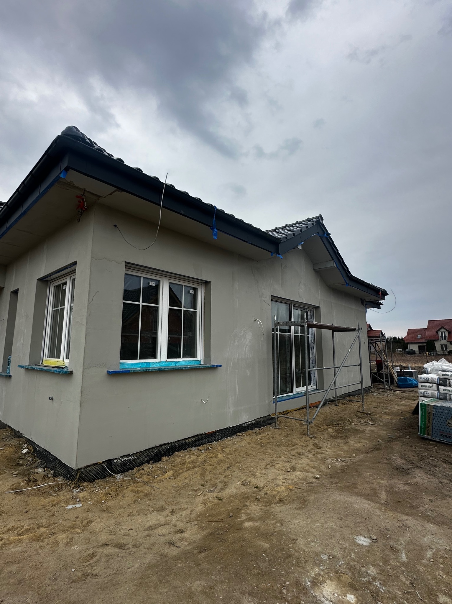 Dom jednorodzinny w trakcie ocieplania elewacji, widoczne rusztowania i materiały budowlane. Szara elewacja, białe okna, pochmurne niebo w tle.