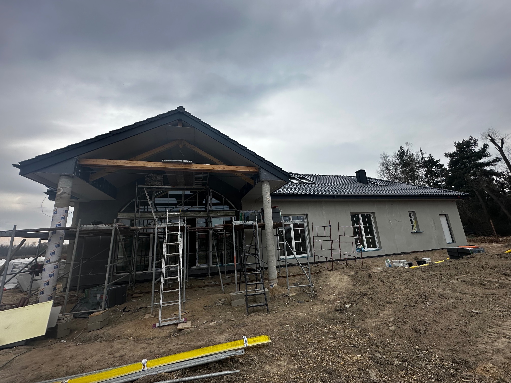 Dom w trakcie budowy z szarym tynkiem, widoczne rusztowania i elementy konstrukcyjne dachu. Pochmurne niebo w tle. Ujęcie z dołu.