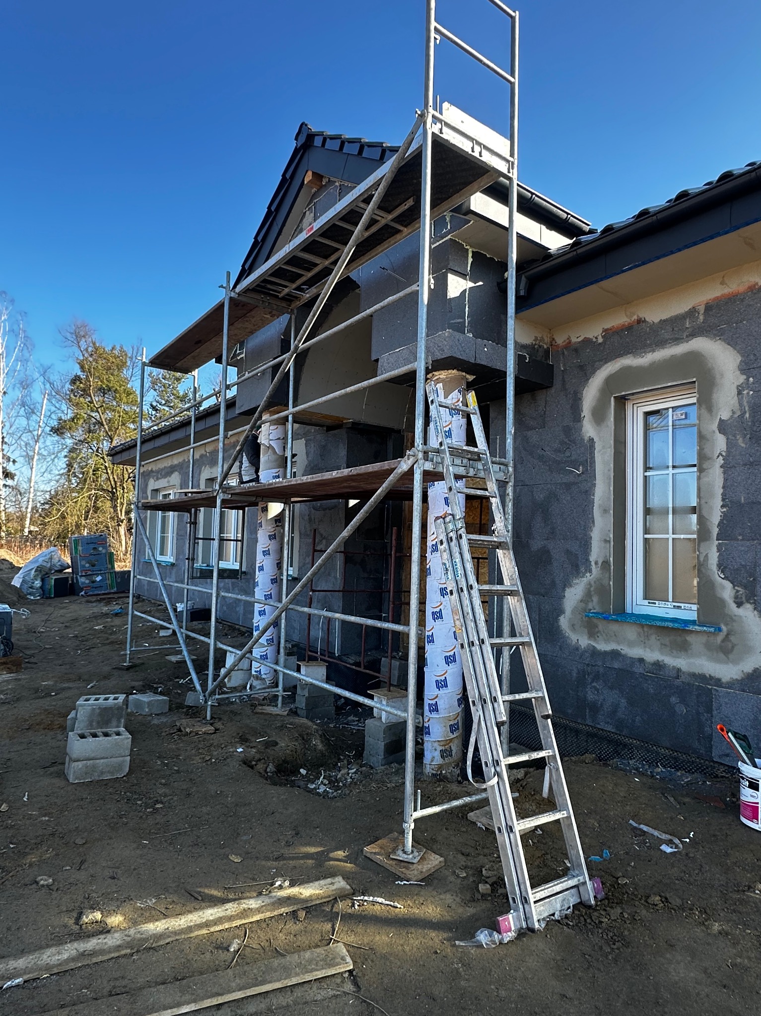 Dom w trakcie ocieplania ścian zewnętrznych, widoczne rusztowania i materiały izolacyjne. Okno z nową ramą. Prace w toku, słoneczny dzień.