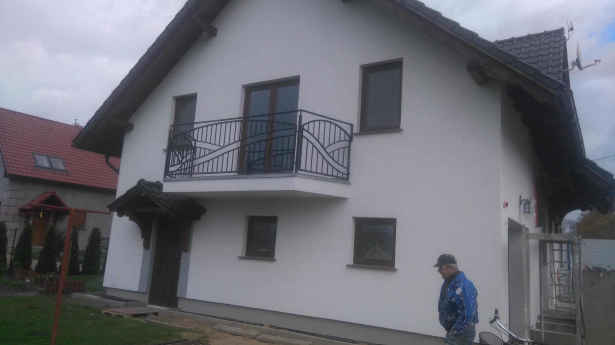 Biały dom jednorodzinny po remoncie elewacji z balkonem o zdobionej, czarnej balustradzie. Na pierwszym planie mężczyzna w roboczym ubraniu i rower.