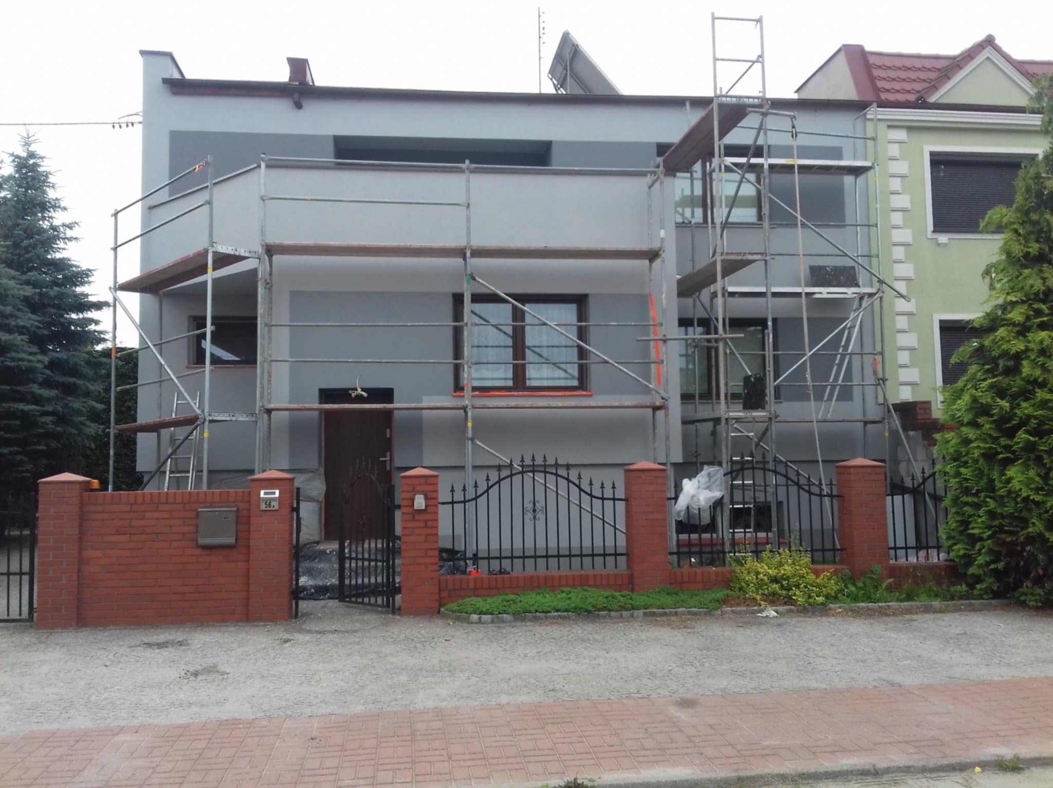 Dom jednorodzinny w trakcie malowania elewacji, otoczony rusztowaniami. Widoczny fragment ogrodzenia z cegły i metalu. Prace w toku, szara kolorystyka budynku.