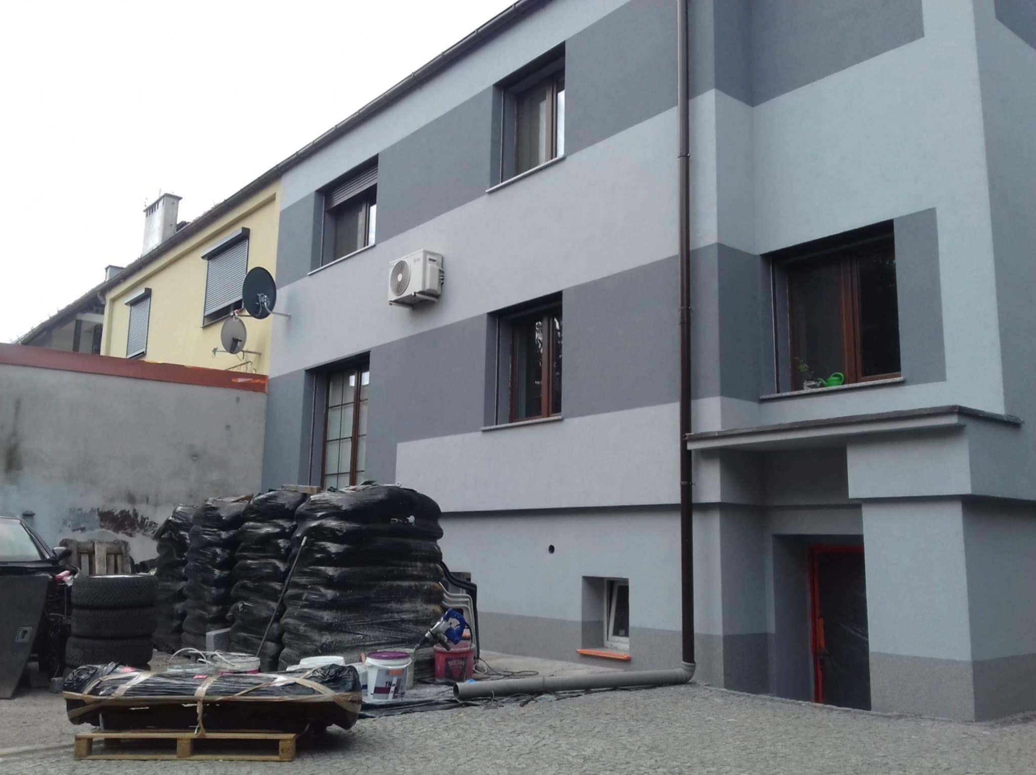 Ocieplony budynek z szarą elewacją w pasy, widoczne materiały budowlane i narzędzia. Detal: klimatyzator na ścianie.