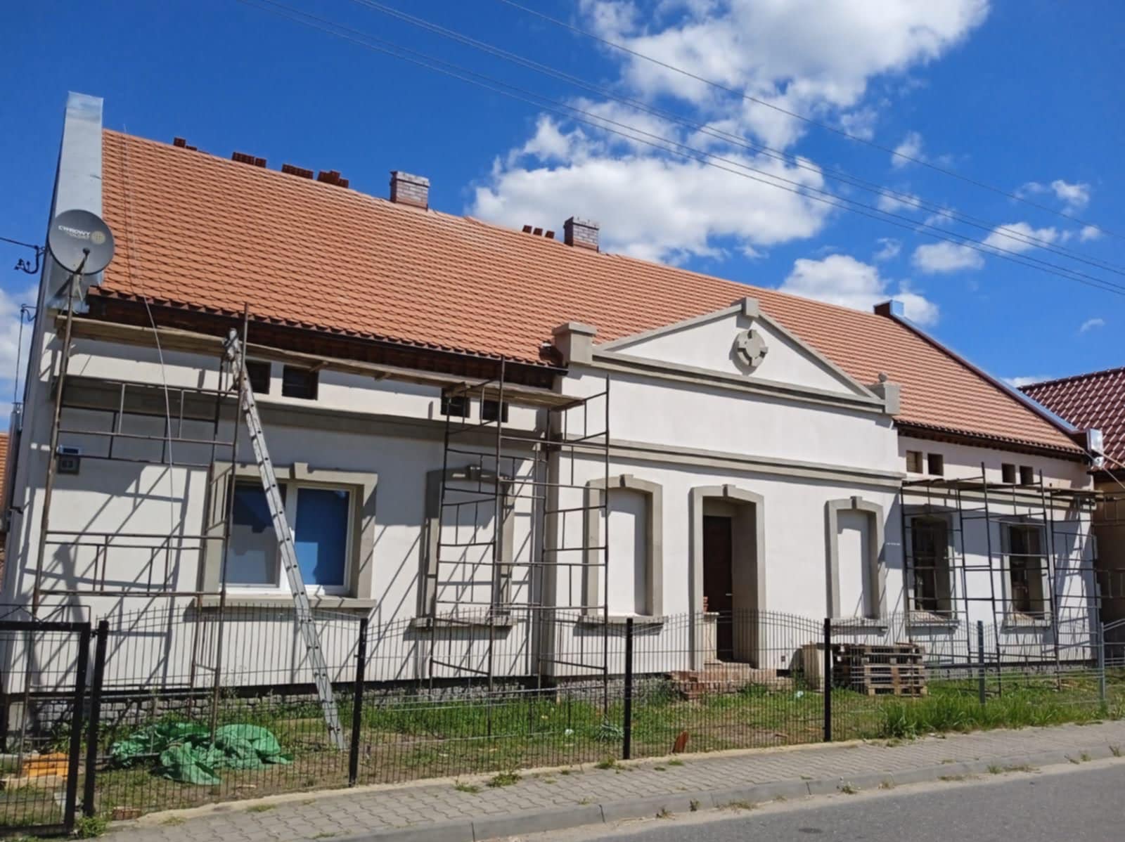 Biały, parterowy dom z czerwoną dachówką w trakcie renowacji elewacji. Widoczne rusztowania, drabina i fragment chodnika. Błękitne niebo z chmurami w tle.